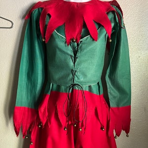 K&ouml;nnte beinhalten: Ein gr&uuml;nes und rotes Elfenkost&uuml;m, das auf einer Schaufensterpuppe ausgestellt ist. Das Kost&uuml;m besteht aus einer gr&uuml;nen Tunika mit rotem Kragen und Manschetten sowie einer roten Hose. Goldene Gl&ouml;ckchen sind an den Manschetten und der Hose befestigt. Die Tunika hat einen schwarzen Schn&uuml;rverschluss.
