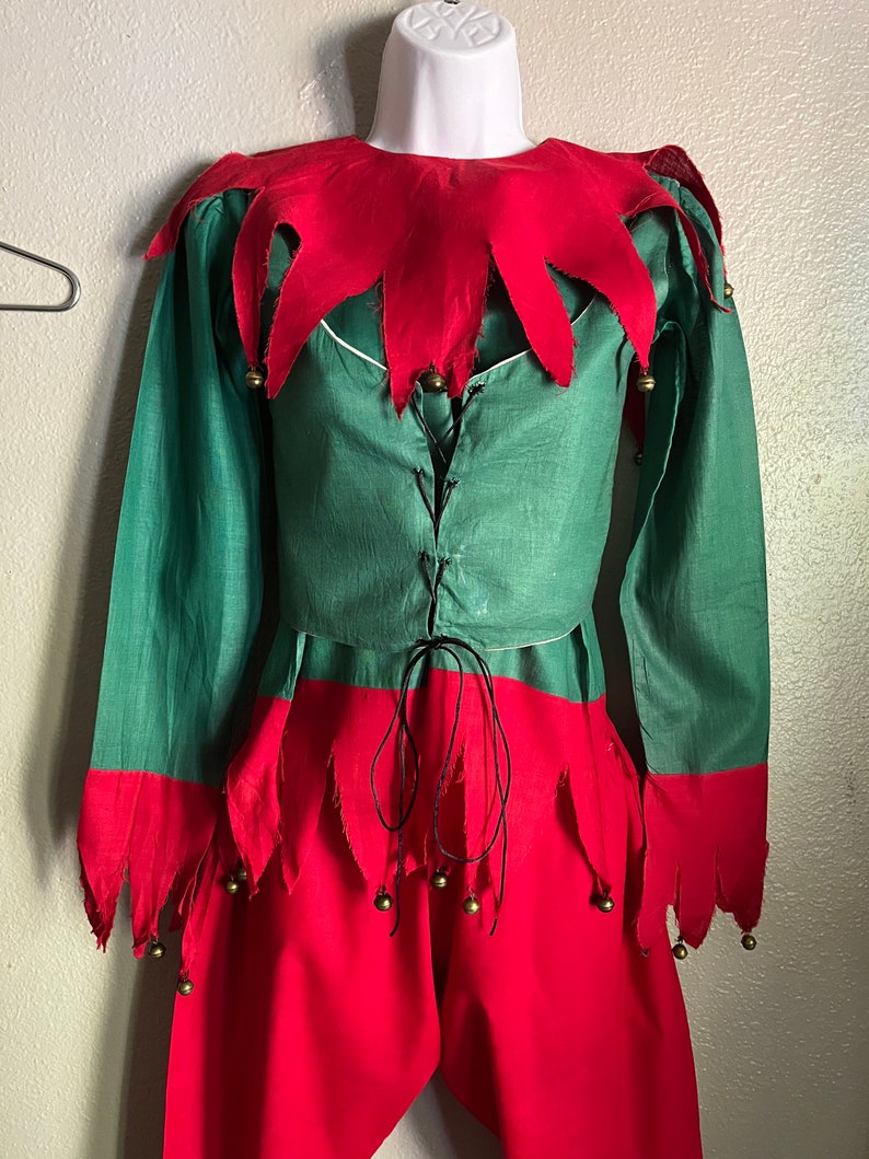 K&ouml;nnte beinhalten: Ein gr&uuml;nes und rotes Elfenkost&uuml;m, das auf einer Schaufensterpuppe ausgestellt ist. Das Kost&uuml;m besteht aus einer gr&uuml;nen Tunika mit rotem Kragen und Manschetten sowie einer roten Hose. Goldene Gl&ouml;ckchen sind an den Manschetten und der Hose befestigt. Die Tunika hat einen schwarzen Schn&uuml;rverschluss.