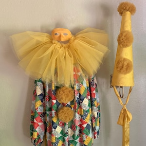 May include: A clown costume featuring a long-sleeved garment with a multi-colored patchwork pattern of small geometric shapes. It includes a large yellow tulle ruff collar, a yellow mask, two yellow pom-poms on the front, and a tall yellow hat.