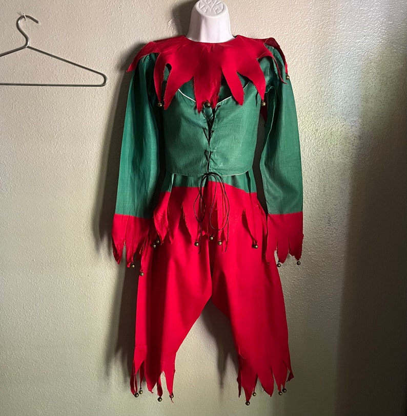 K&ouml;nnte beinhalten: Ein gr&uuml;nes und rotes Elfenkost&uuml;m, das auf einer Schaufensterpuppe ausgestellt ist. Das Oberteil ist gr&uuml;n mit rotem Kragen und Manschetten. Die rote Hose hat gezackte Kanten und kleine Gl&ouml;ckchen. Eine schwarze Schnur schn&uuml;rt die Vorderseite des Oberteils.