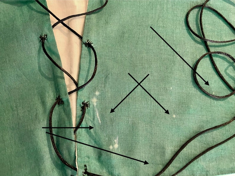 K&ouml;nnte beinhalten: Nahaufnahme eines t&uuml;rkisfarbenen Stoffes mit schwarzer Kordelschn&uuml;rung. Der Stoff hat eine einfarbige t&uuml;rkisfarbene Farbe mit sichtbarer Textur. Die schwarze Kordel ist durch &Ouml;sen an den R&auml;ndern gef&auml;delt und erzeugt ein dekoratives Muster. Der Hintergrund ist ein wei&szlig;er Stoff.