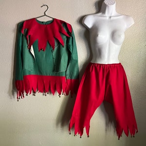 K&ouml;nnte beinhalten: Ein gr&uuml;nes und rotes Elfenkost&uuml;m. Das lang&auml;rmlige Oberteil hat einen roten Kragen und Saum mit kleinen Gl&ouml;ckchen. Die rote Hose hat einen passenden Saum. Das Kost&uuml;m wird auf einem Kleiderb&uuml;gel und einer Schaufensterpuppe pr&auml;sentiert.