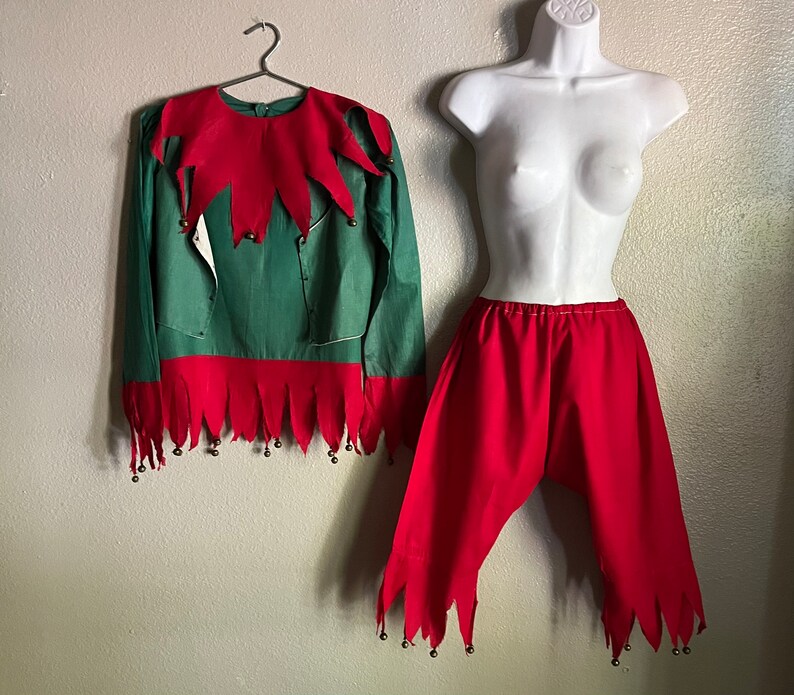 K&ouml;nnte beinhalten: Ein gr&uuml;nes und rotes Elfenkost&uuml;m. Das lang&auml;rmlige Oberteil hat einen roten Kragen und Saum mit kleinen Gl&ouml;ckchen. Die rote Hose hat einen passenden Saum. Das Kost&uuml;m wird auf einem Kleiderb&uuml;gel und einer Schaufensterpuppe pr&auml;sentiert.