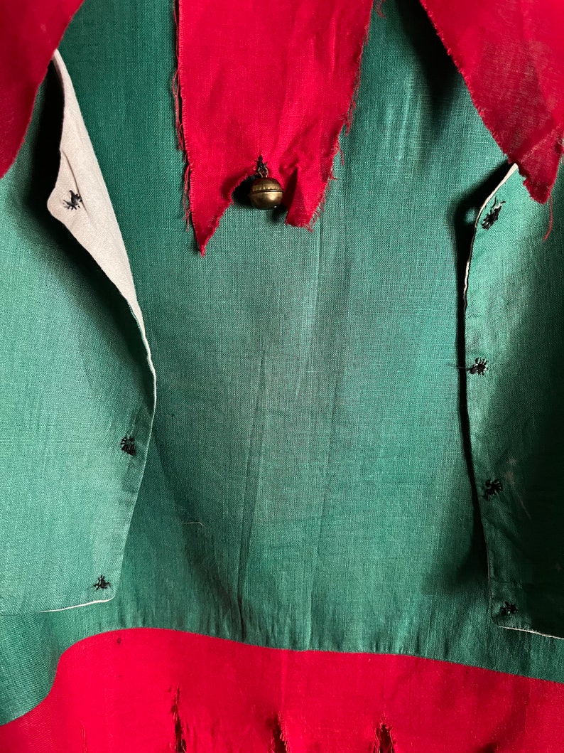 K&ouml;nnte beinhalten: Ein gr&uuml;nes und rotes Weihnachtselfenkost&uuml;m. Das Kost&uuml;m hat eine gr&uuml;ne Tunika mit rotem Kragen und Saum. Eine kleine goldene Glocke h&auml;ngt am Kragen. Die Tunika hat schwarze Kn&ouml;pfe und wei&szlig;e Verzierungen.