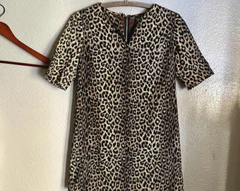 May include: A short-sleeved dress with a leopard print pattern. The dress is hanging on a wooden hanger. The dress has a V-neckline and a zipper closure. The fabric has a tan background with black spots.