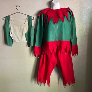 K&ouml;nnte beinhalten: Ein gr&uuml;nes und rotes Elfenkost&uuml;m mit passender Weste. Das lang&auml;rmlige Hemd und die Hose sind rot mit gr&uuml;nen Akzenten und goldenen Gl&ouml;ckchen. Die Weste ist gr&uuml;n mit einer cremefarbenen Vorderseite. Das Kost&uuml;m wird auf einer Schaufensterpuppe pr&auml;sentiert.