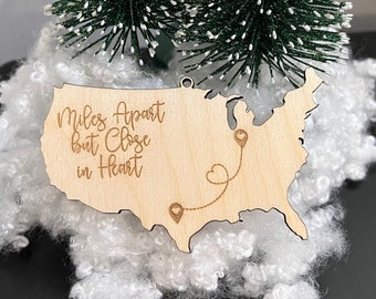 May include: Wooden ornament shaped like the United States with a heart-shaped string connecting two points on the map. The ornament reads "Miles Apart but Close in Heart".
