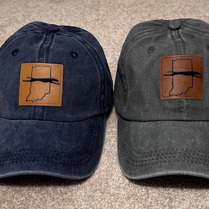 May include: Two baseball caps, one navy blue and one gray, with a distressed finish. Each cap features a brown leather patch with the outline of Indiana and a horizontal line.