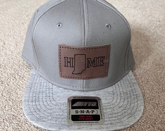 May include: A gray snapback hat with a gray brim. A brown leather patch on the front displays the word "HOME" with the outline of the state of Indiana. The hat has a snap closure and a tag that reads "ONE SIZE FITS MOST".