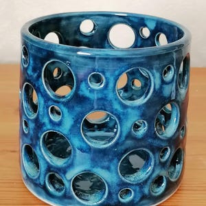 May include: A cylindrical ceramic vessel featuring a glossy, deep blue glaze with a mottled finish. The exterior wall is perforated with numerous circular cutouts of varying sizes, creating an open, airy structure throughout the piece.