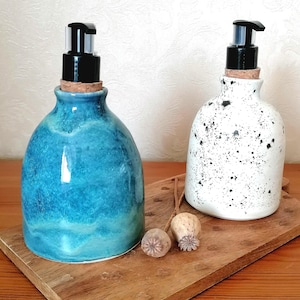 May include: Two ceramic soap dispensers with black pumps and cork stoppers. One is a glossy turquoise, the other white with black speckles. They sit on a wooden tray with dried seed pods.