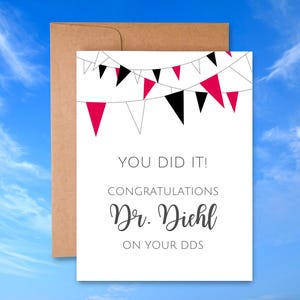 May include: A white greeting card with the text "YOU DID IT! CONGRATULATIONS Dr. Diehl ON YOUR DDS" is displayed. The card features a banner of black and red pennants. The card is inside a brown envelope.