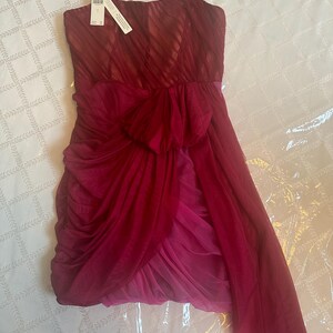 K&ouml;nnte beinhalten: Ein tr&auml;gerloses, knielanges Kleid in Rot- und Rosat&ouml;nen. Das Oberteil hat ein strukturiertes, diagonales Streifenmuster, und der Rock hat einen mehrlagigen, drapierten Stoff mit einer Schleife in der Taille. D&uuml;nne Spaghettitr&auml;ger und ein Etikett sind angebracht.