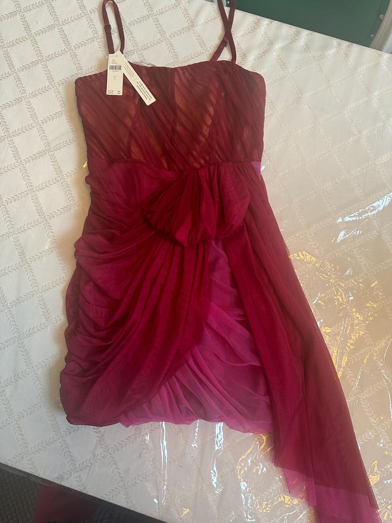 K&ouml;nnte beinhalten: Ein tr&auml;gerloses, knielanges Kleid in Rot- und Rosat&ouml;nen. Das Oberteil hat ein strukturiertes, diagonales Streifenmuster, und der Rock hat einen mehrlagigen, drapierten Stoff mit einer Schleife in der Taille. D&uuml;nne Spaghettitr&auml;ger und ein Etikett sind angebracht.