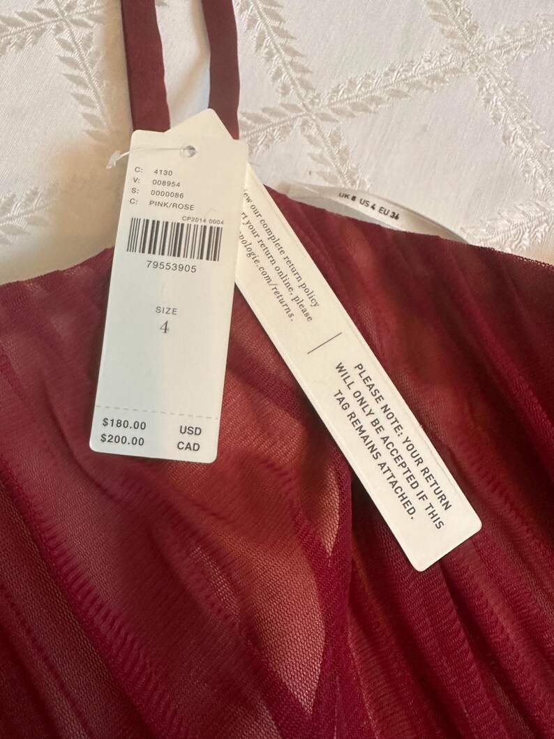K&ouml;nnte beinhalten: Ein bordeauxfarbenes Kleid mit einem transparenten Overlay und einem angebrachten Etikett. Das Etikett gibt die Kleidergr&ouml;&szlig;e 4 an und enth&auml;lt den Text "PLEASE NOTE: YOUR RETURN WILL ONLY BE ACCEPTED IF THIS TAG REMAINS ATTACHED."