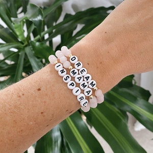 Peut inclure: Quatre bracelets en perles avec les mots "DREAM", "MAMA", "INSPIRE" et "IRE" dessus. Les bracelets sont faits de perles roses et d'accents dorés.