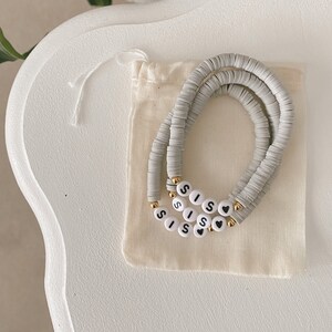May include: A set of two grey beaded bracelets with the word "SISS" spelled out in black letters on white beads. The bracelets are in a small white drawstring bag.