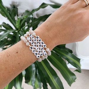 Peut inclure: Un ensemble de cinq bracelets en perles avec les mots "Mama", "Strength", "Inspire", "Mom" et un symbole de cœur. Les bracelets sont faits de perles blanches avec des lettres noires et des accents dorés.