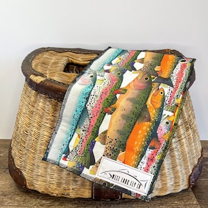 May include: A colorful, patterned cloth featuring illustrations of various fish, resting on a woven wicker basket with a leather trim. The cloth has a black border and a white label with the text "WEST FORK FLY CO."