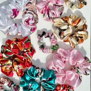 Könnte beinhalten: Eine Sammlung von Satin-Haargummis in verschiedenen Farben und Mustern. Die Haargummis umfassen Weiß mit rosa Blumen, Rosa, Türkis, Rot mit orangefarbenen und gelben Blumen und Creme mit Blumenmustern. Einige sind klein, andere größer.