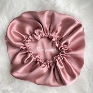 Peut inclure: Un bonnet en satin rose poudré avec un bord élastique froncé. Le bonnet porte l'inscription "Rose poudré" en blanc. Le bonnet est posé sur une surface blanche et duveteuse.