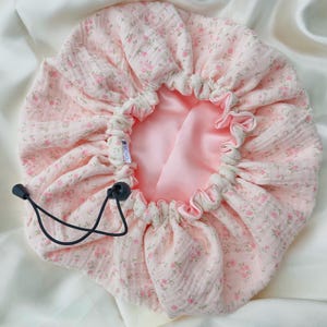 May include: A round hair bonnet featuring a light pink fabric with a small floral pattern on the exterior and a smooth, solid light pink lining. The bonnet includes an adjustable black drawstring with a plastic toggle and a small white label reading SACHOU.