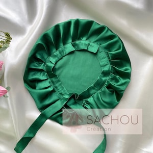 Puede incluir: Un gorro de satén verde esmeralda con borde con volantes y lazos largos. El gorro está diseñado para proteger el cabello y se muestra sobre un fondo de satén blanco. La marca "SACHOU Création" es visible.