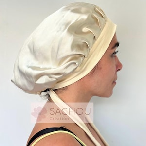 Peut inclure: Un bonnet en satin crème avec un bandeau et des liens assortis. Le bonnet est conçu pour protéger les cheveux et est fabriqué dans un tissu lisse et brillant. La marque "SACHOU Création" est visible.