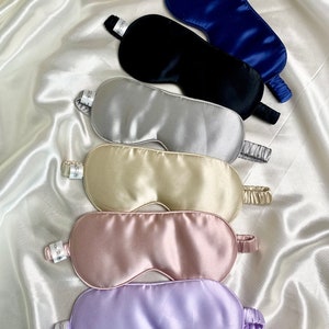 Puede incluir: Varios antifaces de seda en colores como azul marino, negro, plateado, beige, rosa y lavanda. Cada antifaz tiene una cinta elástica y una pequeña etiqueta con texto. Los antifaces están colocados sobre una tela blanca y sedosa.