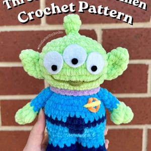 May include: A handmade, green and blue crochet alien toy with three eyes. The alien has a light green head and arms, with a blue body and dark blue pants. The text "Three-Eyed Space Alien Crochet Pattern" is displayed at the top of the image.