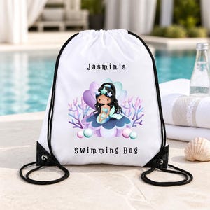 Może przedstawiać: Biały worek ze sznurkiem z czarnym napisem Jasmin's Swimming Bag. Centralny wzór przedstawia rysunkową syrenkę z czarnymi włosami i turkusowym ogonem, siedzącą na fioletowej muszli w otoczeniu koralowców i pereł.