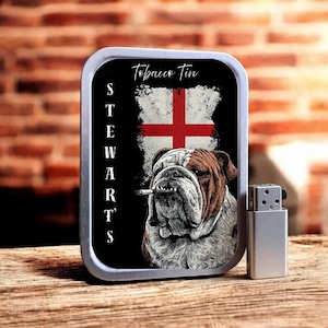 May include: A rectangular metal tobacco tin featuring a bulldog illustration smoking a cigarette, the English flag, and the words "Tobacco Tin" and "Stewarts". A silver lighter is next to the tin.