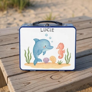 Può includere: Un portapranzo in metallo blu con un pannello frontale bianco con un'illustrazione ad acquerello di un delfino blu, un cavalluccio marino e il nome "Lucie". Il portapranzo ha una maniglia nera ed è appoggiato su una superficie di legno.
