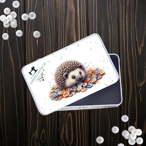 May include: A white metal tin with a hedgehog illustration surrounded by colorful buttons. The tin reads "Grandma's Button Tin" and sits on a dark wooden surface. White buttons are scattered around the tin.