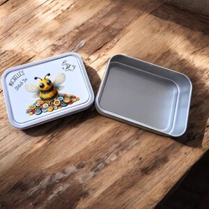 May include: An open, rectangular tin with a lid. The lid has a cartoon bee design, with a yellow and black bee on a pile of buttons. Text on the lid says "Michelle's Stitch Tin" and "Busy as a Bee!"