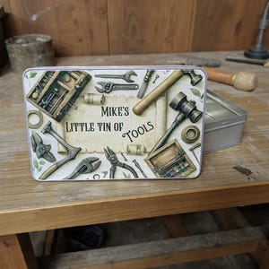 May include: A rectangular tin with a lid, featuring a printed design of various tools. The text "Mike's Little Tin of Tools" is displayed on a scroll. The tools include wrenches, hammers, and pliers, in a vintage style.