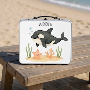 May include: A silver lunchbox with a cartoon orca, starfish, and seaweed design. The name "ABBIE" is printed at the top. The lunchbox is sitting on a wooden picnic table at the beach.
