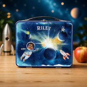 Könnte beinhalten: Eine rechteckige Brotdose aus Metall mit einem dunkelblauen Weltraum-Hintergrund, Planeten, einer Rakete und kleinen Außerirdischen. Der Name RILEY ist in Weiß aufgedruckt, und in der Mitte schwebt ein Kind in einem Astronautenanzug.