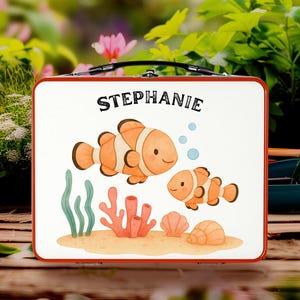 Può includere: Un portapranzo bianco con finiture e manico rossi. Il portapranzo presenta un'illustrazione a cartoni animati di due pesci pagliaccio arancioni, piante marine e il nome "Stephanie" in testo nero.