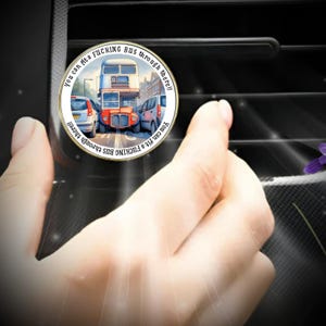 Könnte beinhalten: Ein runder, goldfarbener Auto-Lüftungsclip mit einer Illustration eines Doppeldeckerbusses. Der Text am Rand lautet: "You can fit a FUCKING bus through there!!"