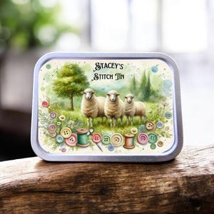 May include: A rectangular metal tin with a watercolour design. It features three sheep in a field, a tree, and sewing notions. The text "STACEY'S STITCH TIN" is printed at the top. The tin is resting on a wooden surface.