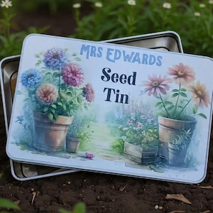 May include: A rectangular seed tin with a floral design. The tin features the text "MRS EDWARDS Seed Tin" in blue and black. The artwork depicts potted flowers in shades of pink, blue, and peach. The tin is set against a blurred garden background.