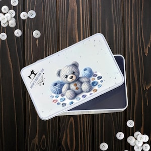 May include: A rectangular white tin with a lid featuring a gray teddy bear, blue yarn, and colorful buttons. The tin is labeled "Mum's Button Tin" with a sewing machine graphic. Scattered white buttons are on a dark wooden surface.