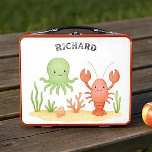 Può includere: Un portapranzo rosso e bianco con il nome "RICHARD" in lettere nere. Il portapranzo presenta un polpo, un'aragosta e piante acquatiche dei cartoni animati su uno sfondo sabbioso. Una mela è accanto al portapranzo.