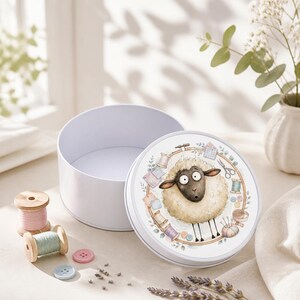 May include: A round white metal storage tin with a lid featuring a cartoon sheep illustration surrounded by sewing supplies like thread spools, scissors, and buttons. Two wooden thread spools and two small buttons are placed next to the tin.
