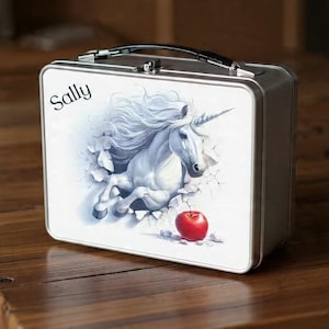 Puede incluir: Una fiambrera de metal plateado con asa y cierre negros. La parte delantera presenta una ilustración de un unicornio blanco rompiendo una pared, una manzana roja y el nombre "Sally" en escritura.
