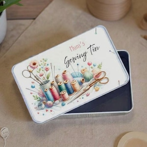 Puede incluir: Una caja de costura rectangular con un diseño de acuarela. La tapa presenta el texto "Mum's Sewing Tin" e ilustraciones de carretes de hilo, tijeras, agujas y flores en tonos rosas, azules y verdes.