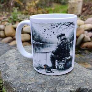 May include: White ceramic mug featuring a black and white illustration of a fisherman by a lake, with a bird in the foreground. The mug has a curved handle and is set against a natural outdoor background.