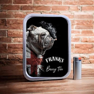May include: A rectangular metal tin featuring a bulldog smoking a cigarette, with the English flag on its chest. The tin is inscribed with "Frank's Baccy Tin". A silver lighter is placed beside the tin.