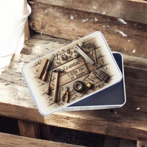May include: A rectangular tin with a blue rim and a lid featuring a wooden plank design. The lid displays various tools, including a hammer, pliers, and a tape roll. The text "Grandad's Little Tin of Tools" is printed on the lid.